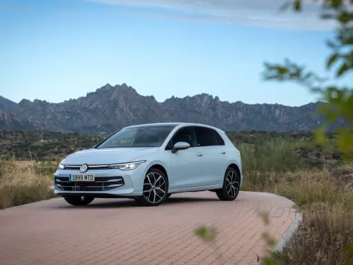 Un coche compacto Volkswagen Golf blanco circula por una carretera de adoquines con montañas rocosas al fondo.