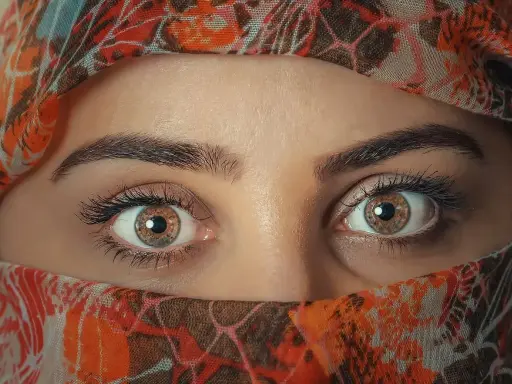 Ojos color miel que miran con curiosidad, enmarcados por un velo con estampado naranja y marrón.