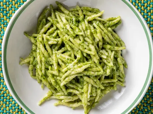 Un piatto di trofie al pesto, pasta fresca condita con un sugo verde brillante, servita in una ciotola bianca su una tovaglietta colorata.
