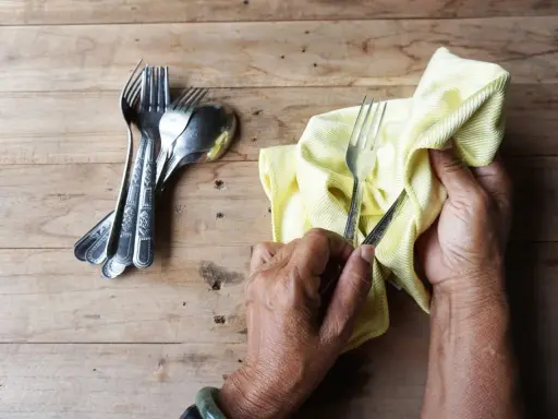Mains polissant des couverts en argent avec un chiffon jaune. Un tas de fourchettes et cuillères est à gauche. Apprenez comment nettoyer l'argenterie.
