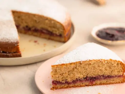 Eine köstliche, schnelle Buchweizentorte mit Marmeladenfüllung und Puderzucker bestäubt. Ein Stück auf einem rosa Teller, der Rest auf einem größeren Teller.