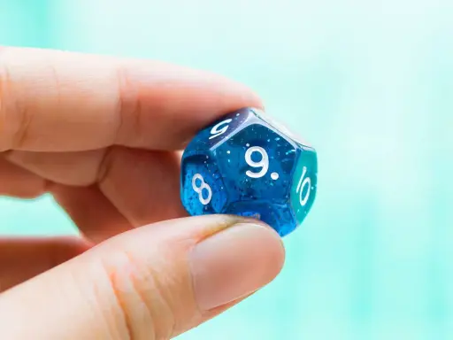 Palec i kciuk trzymają niebieską, dwudziestościenną kostkę z błyszczącymi drobinkami. Widoczna jest cyfra 9, symbolizująca numerologiczną 9.