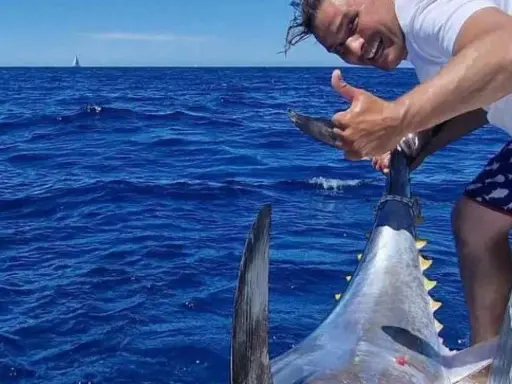 Uomo felice mostra il suo **biggest tuna** appena pescato in mare aperto, con una barca e una vela all'orizzonte.