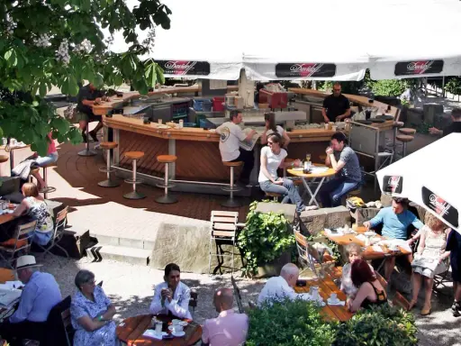 Gäste sitzen an Tischen und an der Theke eines Biergartens unter Sonnenschirmen.