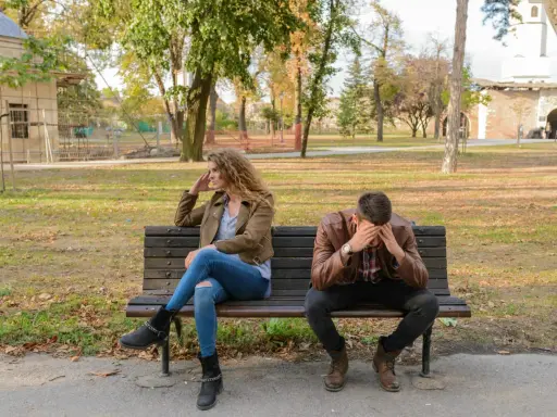 Pareja sentada en un banco, ella mira a un lado, él se tapa la cara. Parecen estar intentando procesar amor y desamor en un parque otoñal.