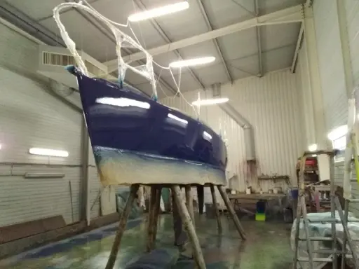 Un bateau bleu profond est en cours de stratification dans un atelier. La coque est soutenue par des tréteaux en bois.
