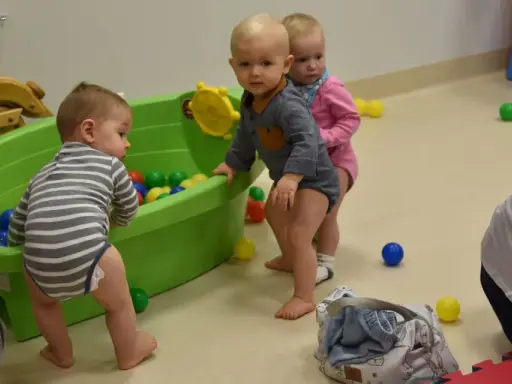 Trzy maluchy, w tym 6 miesięczne dziecko w chodziku, bawią się kolorowymi piłeczkami w zielonej wanience.