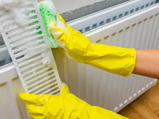 Manos con guantes amarillos limpian la rejilla de un radiador con una esponja y espuma.