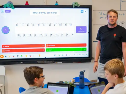 Un insegnante guida una lezione interattiva, simile a Kahoot, con domande su uno schermo touch. I bambini partecipano con entusiasmo.