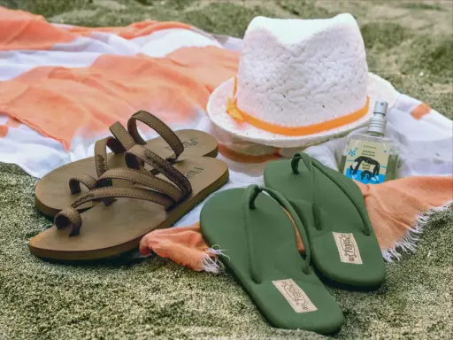Brown sandals vs. green flip flops on a beach towel, with a hat and body mist. A perfect beach day awaits!
