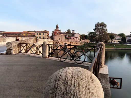 Ponte sul fiume a Borgo San Giuliano, con una bicicletta appoggiata alla ringhiera e palazzi sullo sfondo.