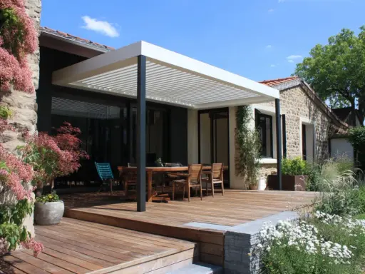 Construction d'une pergola moderne sur une terrasse en bois, avec une table et des chaises, devant une maison en pierre.