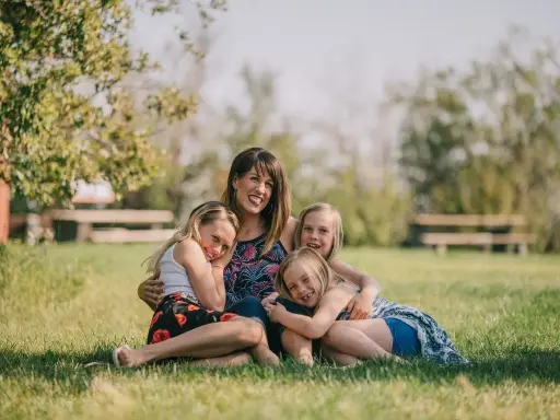 Mama z córkami na trawie, uśmiechnięte i przytulone. Widać, że czują motyle w brzuchu od tej miłości.