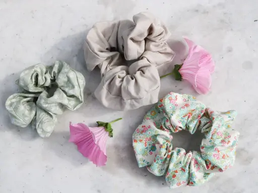 Tre virka scrunchies i olika mönster och färger, omgivna av rosa blommor på en marmorerad yta.