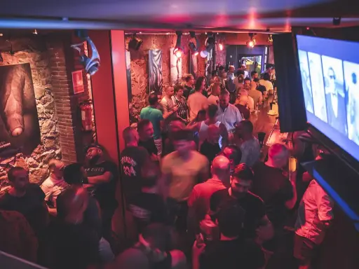 Un locale affollato a Barcellona by night, con uomini che socializzano e bevono. L'atmosfera è vibrante, illuminata da luci rosse e blu.