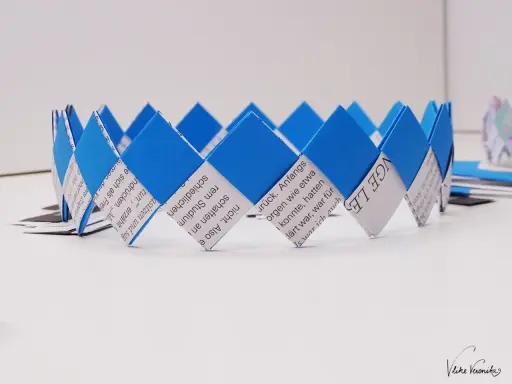 Eine blaue und weiße Papierkrone, die aus gefalteten Papierstreifen gefertigt wurde. Die Streifen sind mit Text bedruckt, was die Krone zu einem besonderen Kunstwerk macht.