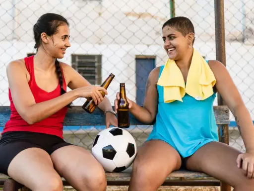 Zwei Frauen mit Fußball und Bierflaschen stoßen an. Nach dem Sport ist vor dem Genuss: sport und alkohol.