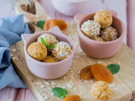 Zwei rosa Schalen gefüllt mit leckeren Energy Balls, bestreut mit Kokosraspeln und Haferflocken. Getrocknete Aprikosen und Minzblätter liegen daneben.