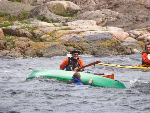 Två män i kajaker nära en klippig kust. En person i en grön kajak verkar ha problem. En diskussion om kanot vs kajak kan uppstå.