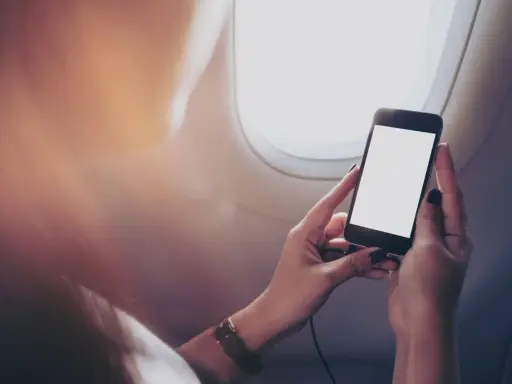 Kobieta trzyma telefon przy oknie w samolocie. Czy w samolocie można korzystać z telefonu? Tak, ale z ograniczeniami.