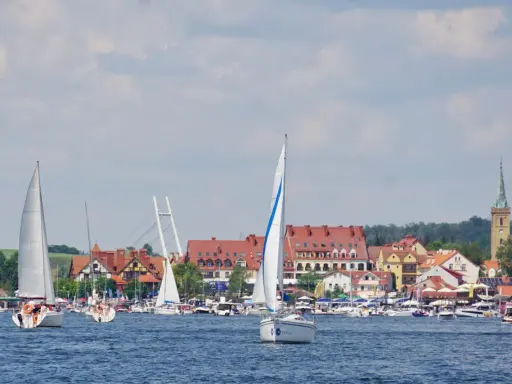 Piękne mazury widoki: żaglówki na jeziorze, miasteczko z czerwonymi dachami i wieżą kościelną w tle.