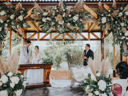 Novios celebran aniversario en altar decorado con flores y pampas. Un oficiante habla mientras la pareja sonríe.