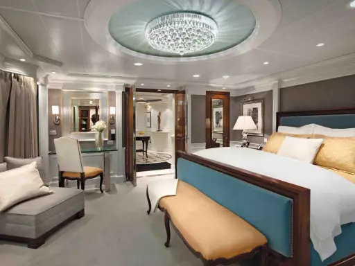 Lussuosa suite crociera con letto king-size, divanetto, lampadario di cristallo e vista su un corridoio elegante.