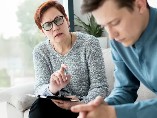 Kobieta w okularach prowadzi terapię odwykową z młodym mężczyzną. Rozmowa o trudnych emocjach i drodze do zdrowia.