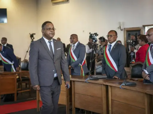 Des hommes en costume, certains portant des écharpes aux couleurs nationales, se tiennent dans une salle. L'un d'eux semble être une persona non grata, marchant à l'écart.