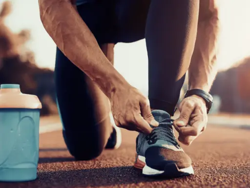 Uomo si allaccia le scarpe da corsa, pronto per correre a stomaco vuoto. Accanto, una borraccia blu.