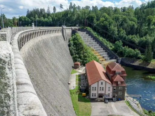 Potężna, kamienna zapora, największa zapora w Polsce, wznosi się nad rzeką. Obok niej budynki elektrowni i zielone wzgórza.