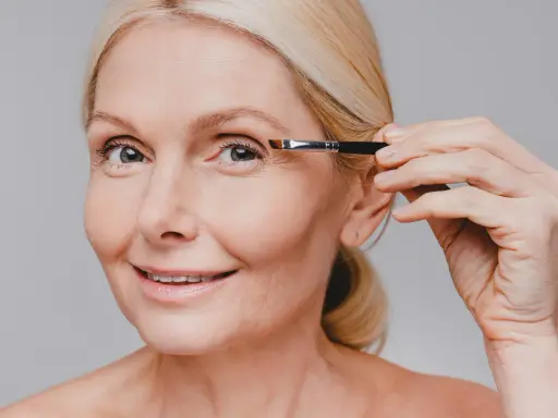 Une femme blonde applique du maquillage sur son œil, son sourcil blanc est visible.