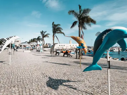 Bunte Delfin-Skulpturen säumen die Promenade in Setúbal, Portugal, unter blauem Himmel und Palmen.