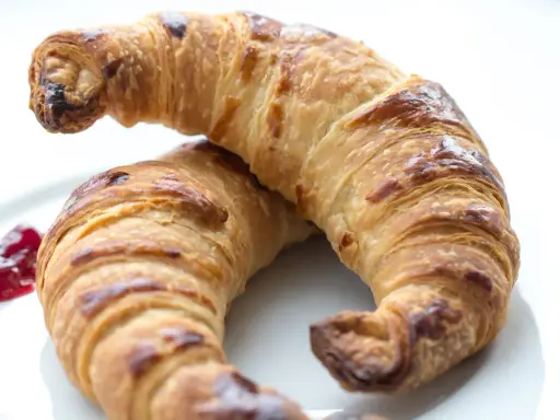 Två gyllene croissants med vaniljfyllning ligger på en vit tallrik. En klick hallonsylt bredvid.