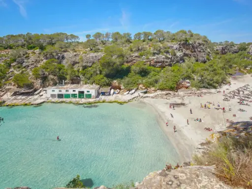 Türkisfarbenes Wasser und ein sonniger Sandstrand in Cala Pi, umgeben von grünen Hügeln. Menschen entspannen sich unter Sonnenschirmen.