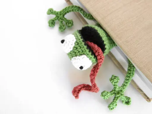 Marcador de libro con ranas amigurumi, verde y negro, con lengua roja, asomando de un libro beige.