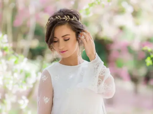Delikatna tiara na głowę zdobi brunetkę w białej sukni z koronkowymi rękawami, otoczoną kwitnącymi drzewami.