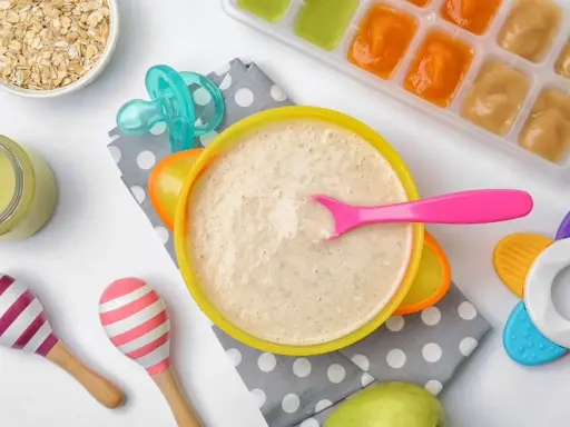 Un tazón de papilla de cereales con una cuchara rosa, junto a avena, purés de frutas y juguetes para bebés.