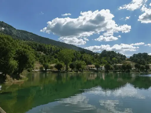 Il lago Sirino, con le sue acque verdi e calme, riflette le case e le colline boscose sotto un cielo azzurro con nuvole bianche.