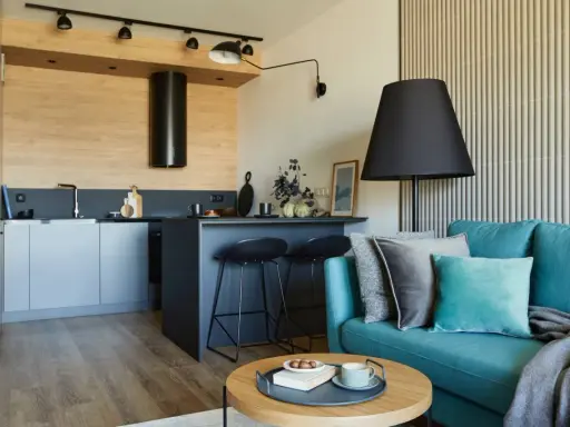 Nowoczesny salon z aneksem kuchennym 22m2. Turkusowa kanapa, czarne hokery przy wyspie, drewniane akcenty i stylowe oświetlenie tworzą przytulną przestrzeń.