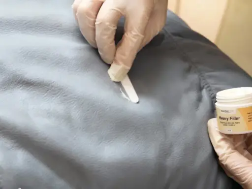 Händer med handskar applicerar filler för att laga reva i tyg. En burk med "Heavy Filler" syns.