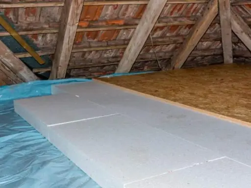Aislamiento de suelo en una casa antigua con paneles de poliestireno sobre plástico azul, bajo vigas de madera y tejas.
