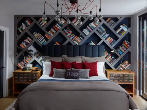 Camera da letto con un'imponente parete di librerie stravaganti a rombi, piene di libri colorati. Un letto accogliente con cuscini rossi e grigi.