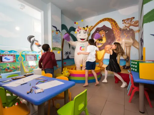 Bambini giocano con un personaggio in costume da ippopotamo in una stanza colorata, un'area giochi per famiglie al Mokambo Shore Easy Family Hotel.