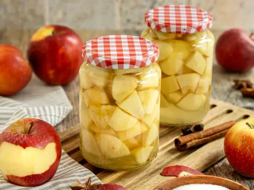 Zwei Gläser mit Apfelstücken, bereit zum Apfel einkochen. Frische Äpfel, Zimtstangen und Sternanis liegen daneben.