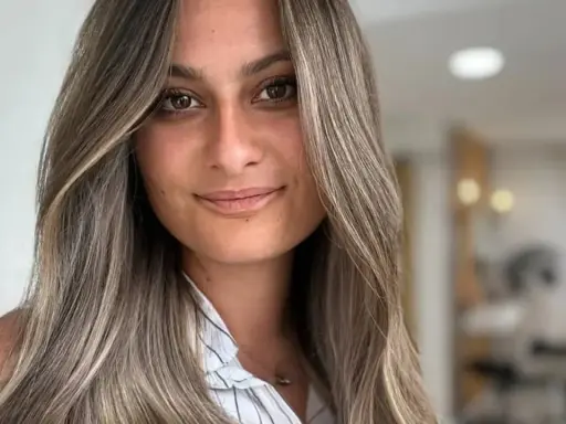 Frau mit langen, welligen Haaren in Balayage-Optik, aschblond mit helleren Strähnen. Ihr Blick ist warm und einladend.