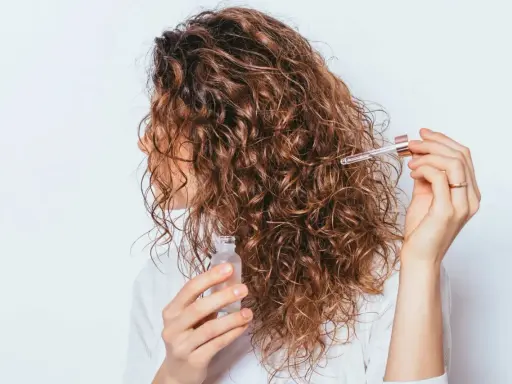 Une femme applique une huile capillaire sur ses cheveux frisottis sur le dessus. Ses boucles brunes brillent sous la lumière.