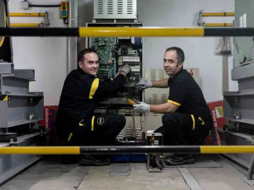 Técnicos realizan mantenimiento de elevadores en un taller, revisando la electrónica y cableado.