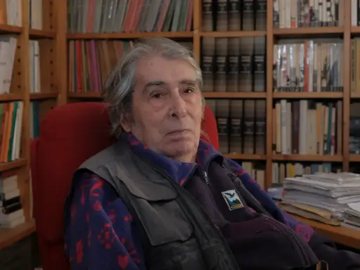Ferdinando Camon, con i suoi capelli grigi e lo sguardo pensieroso, siede tra scaffali pieni di libri, un ritratto di un intellettuale immerso nel suo mondo.
