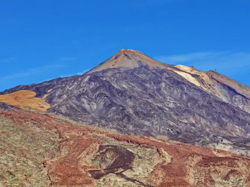 Majestätischer Vulkan im Teide Nationalpark unter blauem Himmel. Die Hänge zeigen verschiedene Gesteinsschichten und eine sandfarbene Fläche.